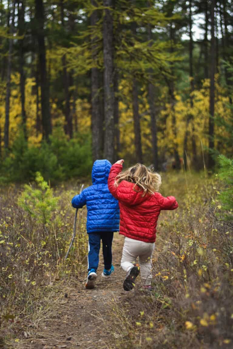 A boy and a girl in a red and blue jacket run along a hiking trail