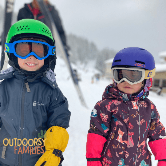 image of two toddlers ready to ski. Wearing helmets, goggles, snowsuits, and mittens.